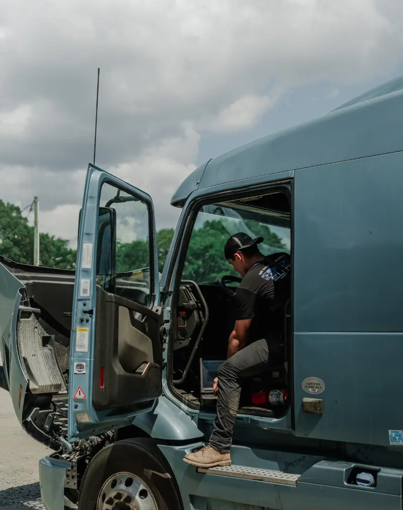 A man in a cap stepping into the open cab of a blue-gray semi truck, with a laptop on the cab floor and the hood tilted open